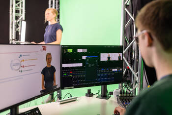 Blick in eine laufende Videoaufzeichnung. Eine Person im Hintergrund steht vor einem Greenscreen, eine andere Person im Vordergrund kontrolliert die Aufnahmen am Bildschirm Blick in eine laufende Videoaufzeichnung. Eine Person im Hintergrund steht vor einem Greenscreen, eine andere Person im Vordergrund kontrolliert die Aufnahmen am Bildschirm